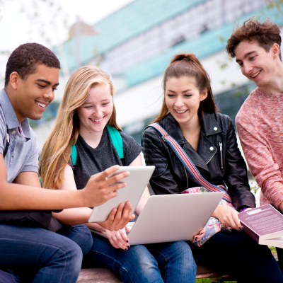 Four diverse students engaged in a collaborative activity outdoors, using a tablet and a laptop.